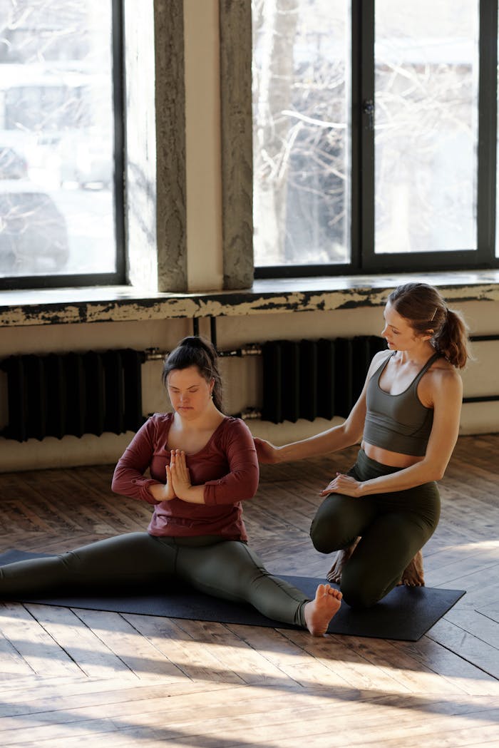 menu-01 A yoga class featuring a personal trainer assisting a woman in a stretching pose indoors.